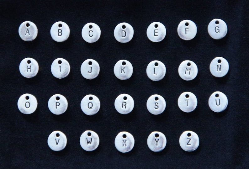 May include: A set of 26 silver metal alphabet letter charms. Each charm is round and has a single letter engraved on it. The letters are in uppercase and are arranged in alphabetical order.