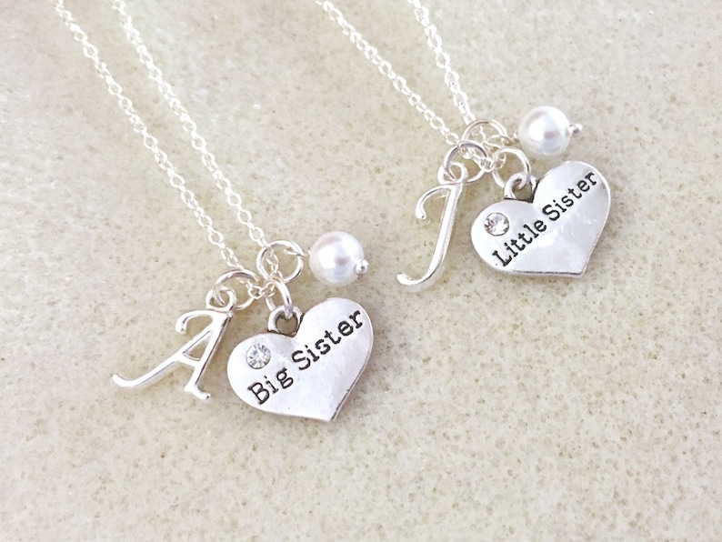 May include: Two silver necklaces with heart pendants. One pendant says "Big Sister" and the other says "Little Sister". Each necklace has a pearl charm and a letter charm.