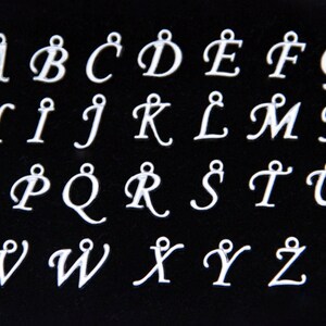 May include: A set of 26 silver-colored metal alphabet charms, each featuring a cursive letter from A to Z. The charms are arranged in three rows on a black background.
