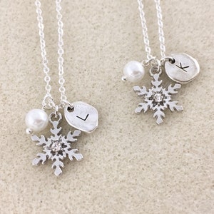 K&ouml;nnte beinhalten: Zwei silberne Halsketten mit Schneeflockenanh&auml;ngern, jeweils mit einer Perle und einem personalisierten Initialanh&auml;nger. Die Initialen sind "K" und "L".