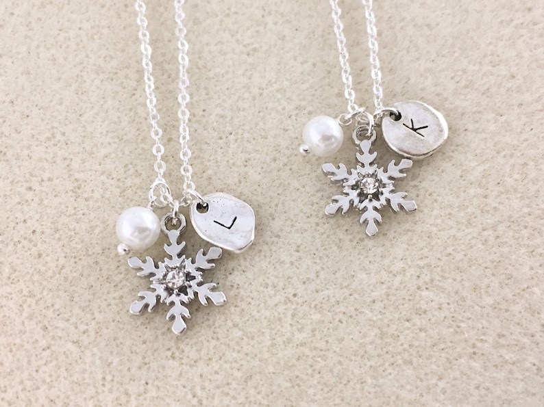 K&ouml;nnte beinhalten: Zwei silberne Halsketten mit Schneeflockenanh&auml;ngern, jeweils mit einer Perle und einem personalisierten Initialanh&auml;nger. Die Initialen sind "K" und "L".