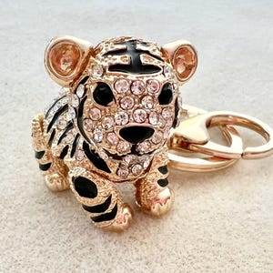 May include: A gold-tone key chain featuring a black and white tiger charm with rhinestones. The tiger charm is detailed with black stripes and has a gold-tone ring attached to the top.