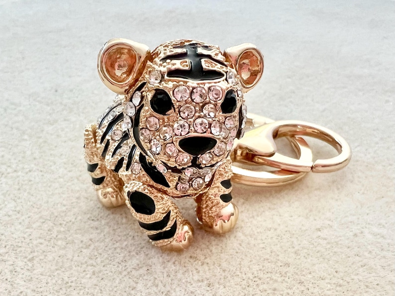 May include: A gold-tone key chain featuring a black and white tiger charm with rhinestones. The tiger charm is detailed with black stripes and has a gold-tone ring attached to the top.