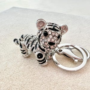 May include: A silver and black tiger keychain with crystal accents. The tiger is sitting on its hind legs with its tail curled around its body. The keychain has a silver ring for attaching to keys.