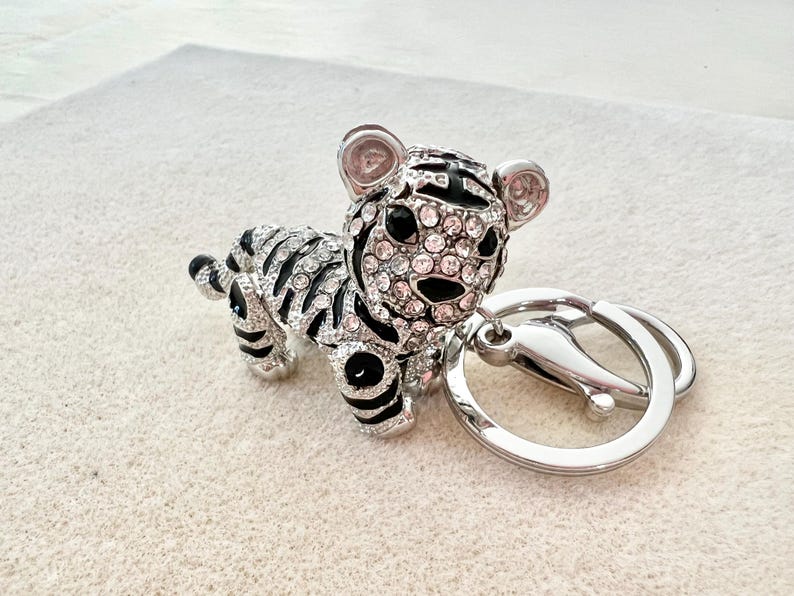 May include: A silver and black tiger keychain with crystal accents. The tiger is sitting on its hind legs with its tail curled around its body. The keychain has a silver ring for attaching to keys.