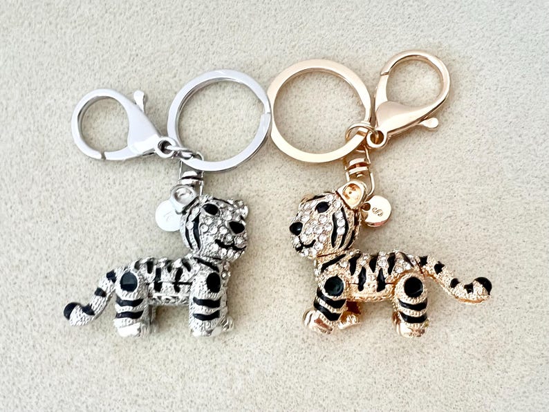 May include: Two silver and gold keychains with tiger charms. The charms are covered in rhinestones and have black stripes.