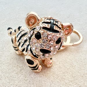 May include: Gold-tone tiger charm with black and white stripes and clear rhinestones. The charm is attached to a gold-tone key ring.