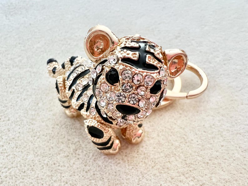May include: Gold-tone tiger charm with black and white stripes and clear rhinestones. The charm is attached to a gold-tone key ring.