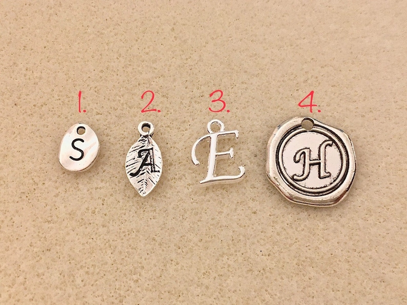 May include: Four silver charms with different designs. The first charm is a round oval with the letter "S" in the center. The second charm is a leaf with the letter "A" in the center. The third charm is the letter "E" in a script font. The fourth charm is a round wax seal with the letter "H" in the center.