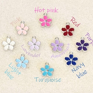Pu&ograve; includere: Assortimento di 10 ciondoli a forma di fiore in vari colori, tra cui rosa acceso, rosso, viola, lavanda, azzurro, turchese e blu navy. Ogni ciondolo &egrave; un fiore in metallo argentato con un centro in smalto colorato.