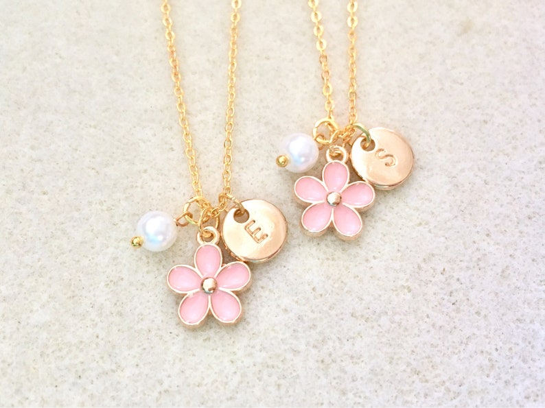Pu&ograve; includere: Due collane d'oro con ciondoli a forma di fiore rosa e dettagli di perle. Ogni collana ha un piccolo ciondolo rotondo con le lettere "LL" e "SS" incise.
