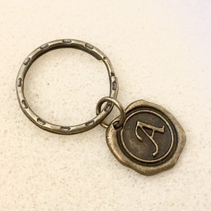May include: A bronze key chain with a round metal ring and a wax seal charm with the letter "A" embossed on it.