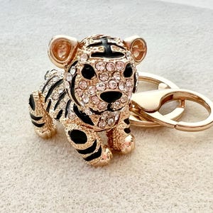 May include: Gold-tone tiger keychain with black and white stripes and clear rhinestones. The tiger is holding a gold-tone ring.
