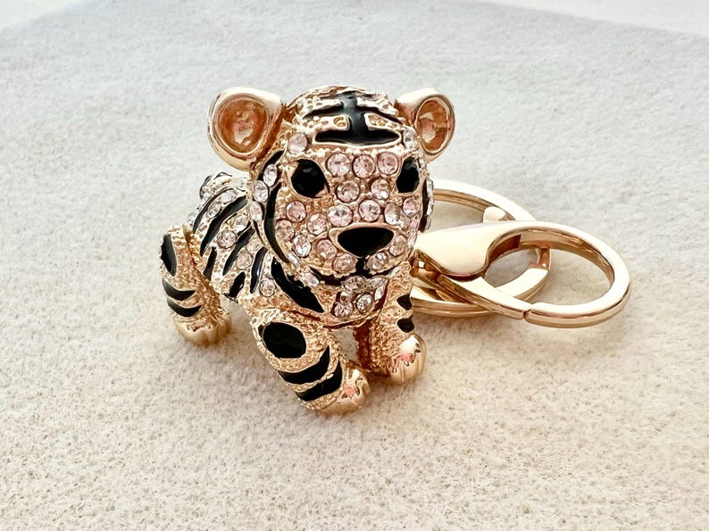 May include: Gold-tone tiger keychain with black and white stripes and clear rhinestones. The tiger is holding a gold-tone ring.