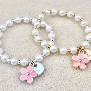 May include: Two pearl bracelets with flower charms and personalized disc charms. The bracelet on the left has a pink flower charm and a disc charm with the letter "LL" on it. The bracelet on the right has a pink flower charm and a disc charm with the letter "A" on it.