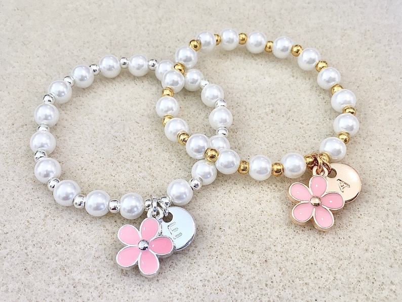 May include: Two pearl bracelets with flower charms and personalized disc charms. The bracelet on the left has a pink flower charm and a disc charm with the letter "LL" on it. The bracelet on the right has a pink flower charm and a disc charm with the letter "A" on it.