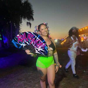 May include: A woman with colorful hair is holding a black and blue fan with the words "Rave Mom" written in pink and white. She is wearing a black bikini top and neon green shorts.