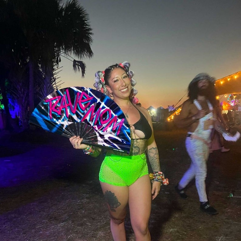 May include: A woman with colorful hair is holding a black and blue fan with the words "Rave Mom" written in pink and white. She is wearing a black bikini top and neon green shorts.