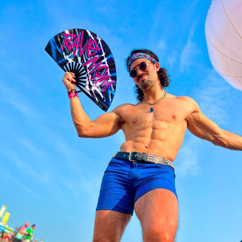 May include: A man wearing blue shorts and a bandana holds a black and blue hand fan with the word "Rave Mom" written on it. He is standing in front of a blue sky.