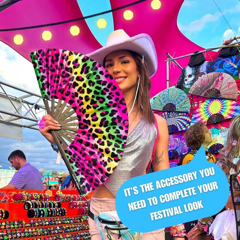 K&ouml;nnte beinhalten: Eine Frau mit einem wei&szlig;en Cowboyhut h&auml;lt einen rosa, gr&uuml;nen und schwarzen Leoparden-Handf&auml;cher. Der F&auml;cher ist aus Stoff und hat einen Holzgriff. Der Text "IT'S THE ACCESSORY YOU NEED TO COMPLETE YOUR FESTIVAL LOOK" steht in einer Sprechblase.