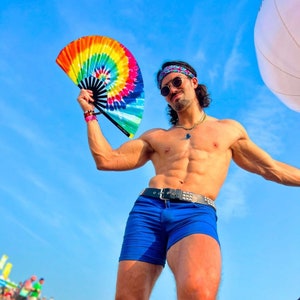 May include: A man wearing a blue bandana and blue shorts holds a rainbow-colored hand fan. He is posing against a blue sky.