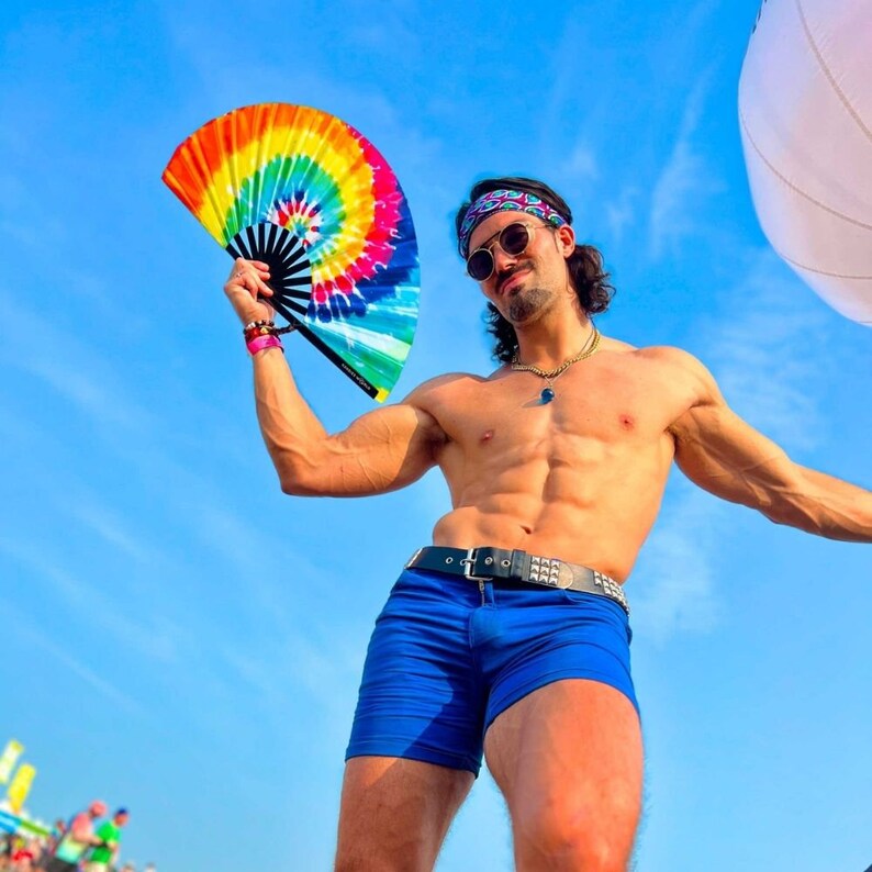 May include: A man wearing a blue bandana and blue shorts holds a rainbow-colored hand fan. He is posing against a blue sky.