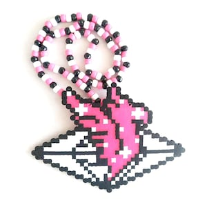 May include: A beaded necklace featuring a pixelated pink heart design. The necklace is made of pink, black, and white beads. The pendant is a heart shape with a black outline and white accents. The necklace is on a white background.