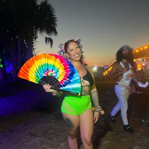 May include: A woman with colorful hair is holding a rainbow tie-dye hand fan. She is wearing a green and black outfit and is standing in front of a green grassy area.
