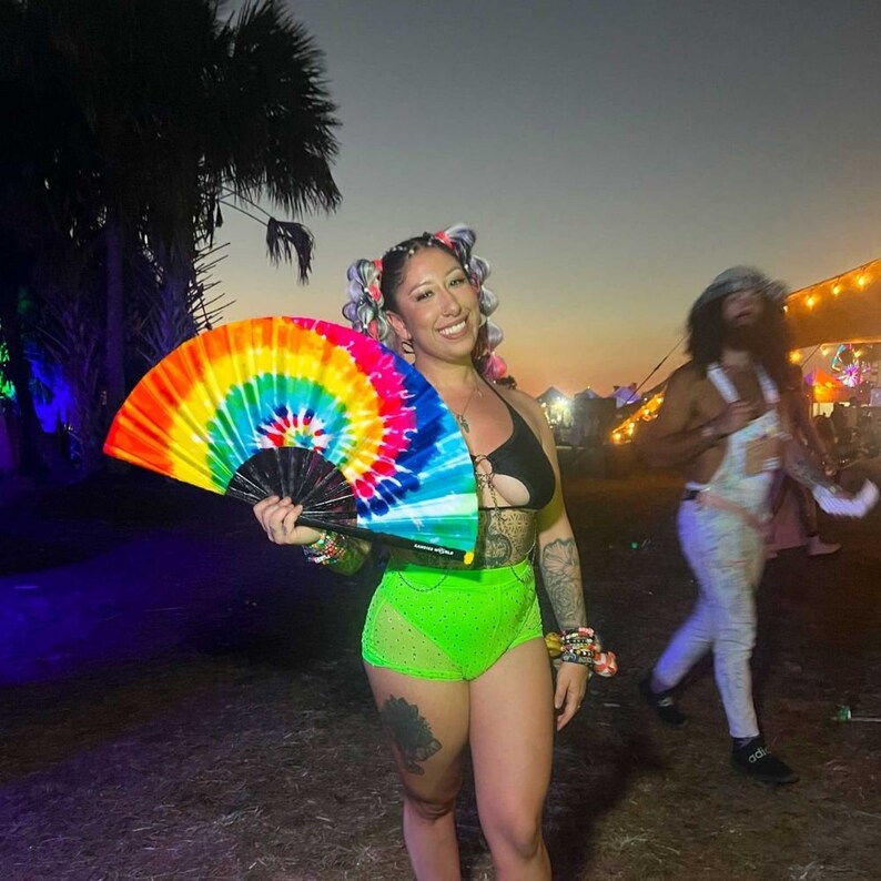 May include: A woman with colorful hair is holding a rainbow tie-dye hand fan. She is wearing a green and black outfit and is standing in front of a green grassy area.