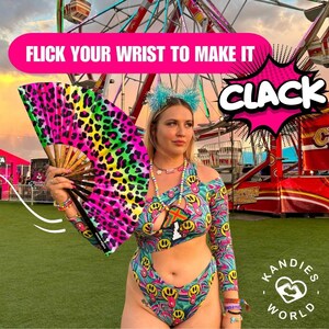 K&ouml;nnte beinhalten: Eine Frau tr&auml;gt einen bunten Badeanzug mit Smiley-Muster und einen Handf&auml;cher mit Leopardenmuster. Der Text "FLICK YOUR WRIST TO MAKE IT CLACK" wird &uuml;ber ihrem Kopf angezeigt. Der Hintergrund ist ein Karnevalssetting mit einem Riesenrad.