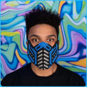May include: A beaded face mask in a geometric design, featuring blue, black, and white beads. The mask covers the lower face and is worn by a person with dark hair and a black shirt. The background has a colorful, swirled pattern.