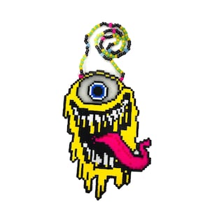 May include: A yellow, pixelated monster pendant with a large eye, open mouth, and pink tongue. The pendant is strung on a beaded necklace with black, yellow, pink, and green beads. The design has a cartoonish, playful aesthetic.
