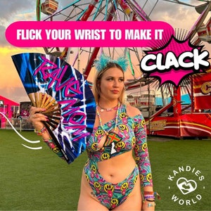 May include: A woman wearing a colorful, psychedelic print bodysuit with a cutout design. She is holding a black and blue hand fan with the words "Rave Mom" written on it. The background is a carnival setting with a Ferris wheel and a sign that says "Clack".