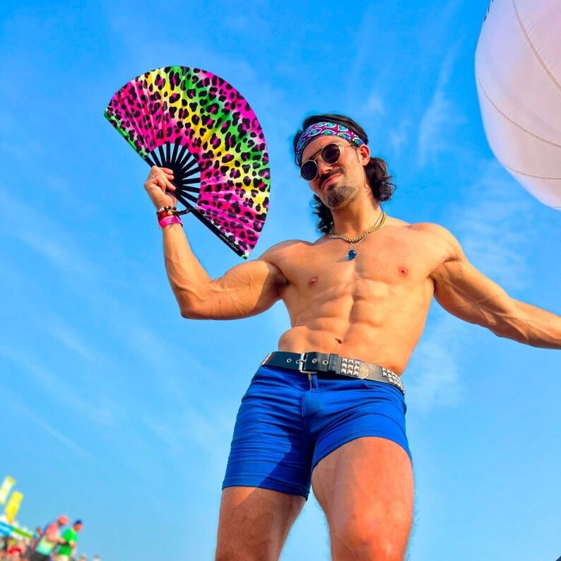 K&ouml;nnte beinhalten: Ein Mann in einer blauen Badehose und einem Handf&auml;cher mit Leopardenmuster. Er tr&auml;gt ein schwarzes Stirnband mit einem bunten Design. Er steht vor einem blauen Himmel.