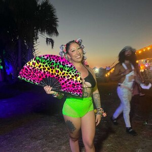 K&ouml;nnte beinhalten: Eine Frau in einem neon-gr&uuml;nen und schwarzen Outfit mit einem Handf&auml;cher mit Leopardenmuster. Sie steht in einem grasbewachsenen Bereich mit B&auml;umen im Hintergrund.