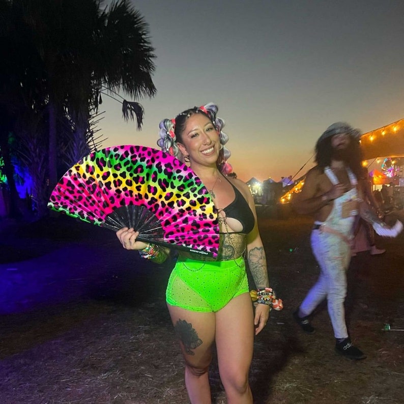 K&ouml;nnte beinhalten: Eine Frau in einem neon-gr&uuml;nen und schwarzen Outfit mit einem Handf&auml;cher mit Leopardenmuster. Sie steht in einem grasbewachsenen Bereich mit B&auml;umen im Hintergrund.