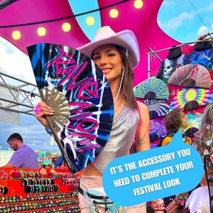 May include: A woman wearing a white cowboy hat holds a blue and pink hand fan with the word "Rave" written in graffiti style. The fan is a festival accessory.