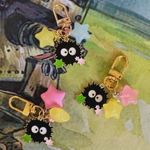 May include: Keychains featuring black, spiky characters with large white eyes, accented by colorful star charms in yellow, pink, and blue. Each keychain has a gold-colored clasp.