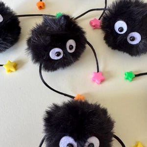 Puede incluir: Una guirnalda caprichosa con pompones negros y esponjosos con ojos blancos y cuentas en forma de estrella de varios colores. Los pompones están conectados por una cuerda negra, creando un elemento decorativo y lúdico.