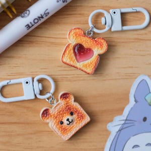May include: Two novelty keychains featuring toast-shaped charms. One charm has a red heart, the other has a bear face. Both charms are attached to white metal clasps, perfect for keys or bags.