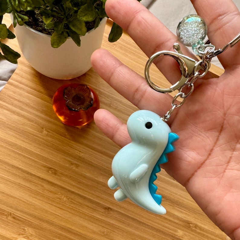 K&ouml;nnte beinhalten: Ein hellblauer Dinosaurier-Schl&uuml;sselanh&auml;nger mit blauem Schwanz und einem silbernen Schl&uuml;sselring. Der Schl&uuml;sselanh&auml;nger hat einen kleinen, durchsichtigen Anh&auml;nger mit wei&szlig;en Glitzerpartikeln im Inneren.