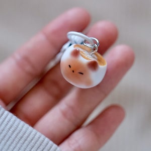 Puede incluir: Un llavero de cerámica blanca con forma de gato enroscado en una bola. El gato tiene marcas marrones y blancas y una pequeña nariz rosa.