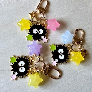 May include: Three gold-toned metal keychains featuring black spiky soot sprite charms with large white eyes. Each keychain is adorned with multiple small, colorful star-shaped charms in shades of yellow, pink, light blue, lime green, and purple.