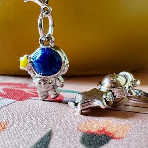 May include: Silver-toned astronaut charm with a blue and yellow helmet. The charm is detailed with a star on the helmet and is attached to a silver chain. A matching astronaut ring is also visible.