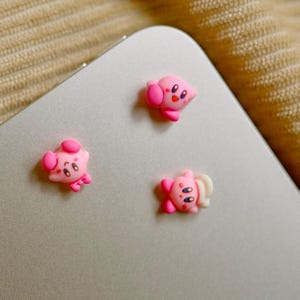 Puede incluir: Tres amuletos rosas de personajes de dibujos animados con ojos negros y mejillas rosadas están dispuestos sobre una superficie plateada. Los amuletos tienen diferentes diseños, uno con una oreja grande y otro con una boca en forma de estrella. Los amuletos son probablemente accesorios decorativos.