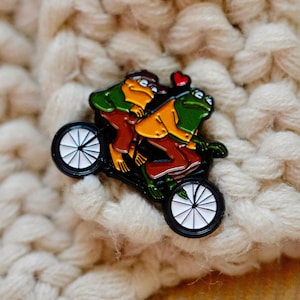 May include: A green and brown frog couple riding a black bicycle with white wheels. The frog in the back is wearing a yellow jacket and brown pants. The frog in the front is wearing a green jacket and brown pants. There is a red heart above the frog's heads.