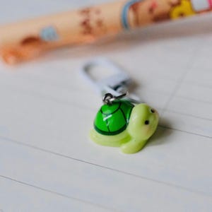 May include: A small, light green turtle charm with a bright green shell and a silver-colored clasp. The turtle has black eyes and is set against a white background. A pen is slightly visible in the background.