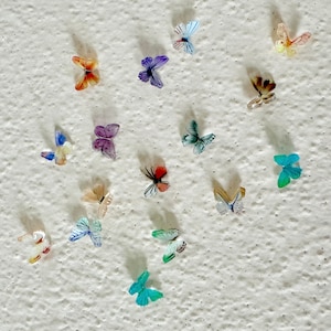 Puede incluir: Una colección de decoraciones de mariposas coloridas en una pared blanca texturizada. Las mariposas están hechas de papel y tienen varios patrones y diseños.