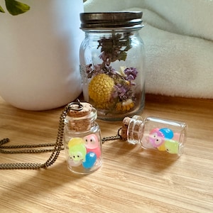 May include: Two small glass jars with cork tops filled with colourful jelly beans. One jar is on a necklace chain and the other is on a table. A larger glass jar with a cork top is filled with dried flowers.