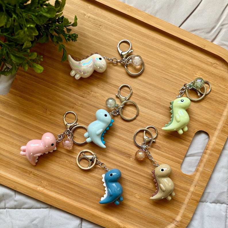K&ouml;nnte beinhalten: Sechs bunte Dinosaurier-Schl&uuml;sselanh&auml;nger mit silbernen Schl&uuml;sselringen und Anh&auml;ngern. Jeder Schl&uuml;sselanh&auml;nger zeigt einen Dinosaurier in einer anderen Farbe mit einem klaren, glitzernden Kugelanh&auml;nger.